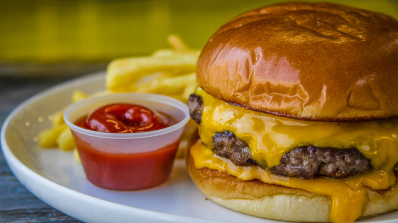 Shiny cheeseburger on a plate with fries and a side cup of ketchup.