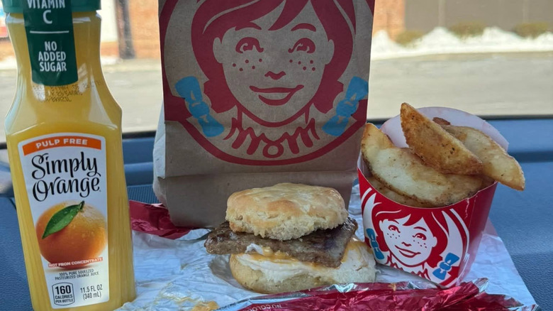 A Wendy's Sausage, Egg & Cheese Biscuit sits next to some orange juice and potato wedges.