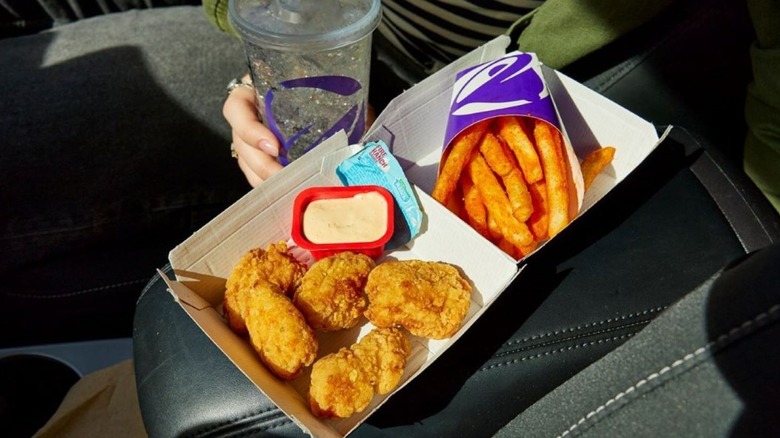 Box of Taco Bell chicken nuggets with an open dipping sauce and fries in a car with a woman's hand holding a soda