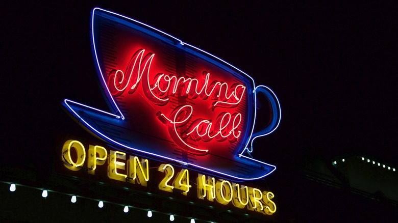 A neon sign of a coffee cup with the words 'Morning Call, Open 24 Hours' written under it