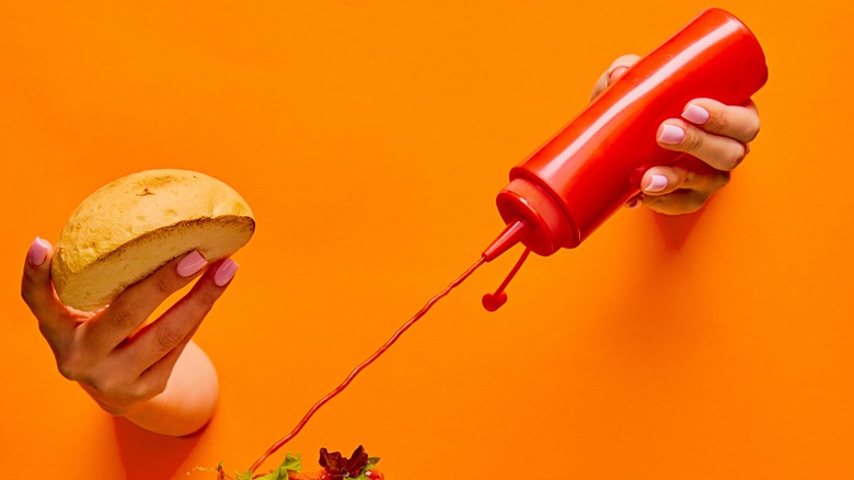 Hands emerge from a wall holding a burger bun and a squeeze bottle of ketchup