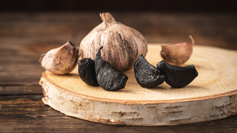 Four peeled cloves of black garlic with two unpeeled cloves and a whole head of garlic behind them sitting on a round surface.