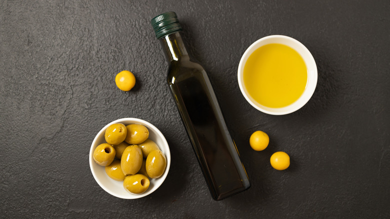 A bottle of olive oil on a dark surface in between a ramekin of green olives and a ramekin of olive oil.