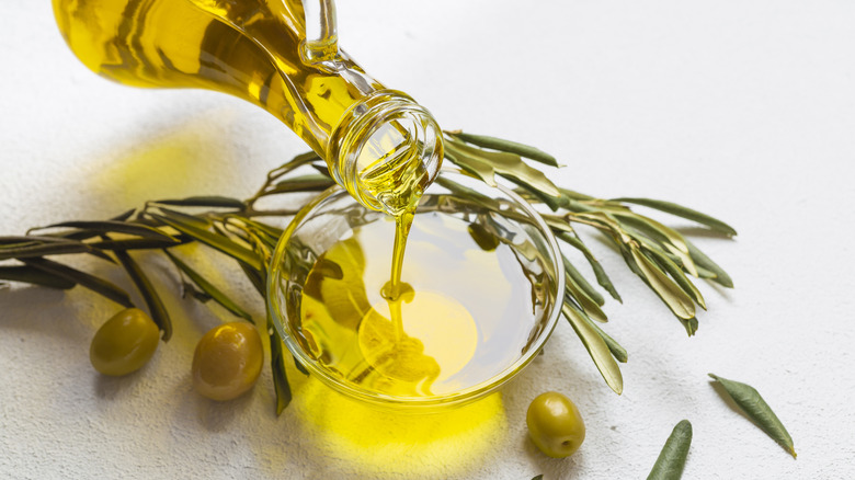 Olive oil being poured into a glass surrounded by green olives and herbs.