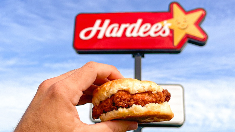 Someone holds up a piece of fried chicken sandwiched in the middle of a biscuit before a Hardee's sign