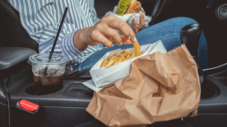 A woman seated in her car lifts a fry with one hand, while holding onto a burger with her other hand