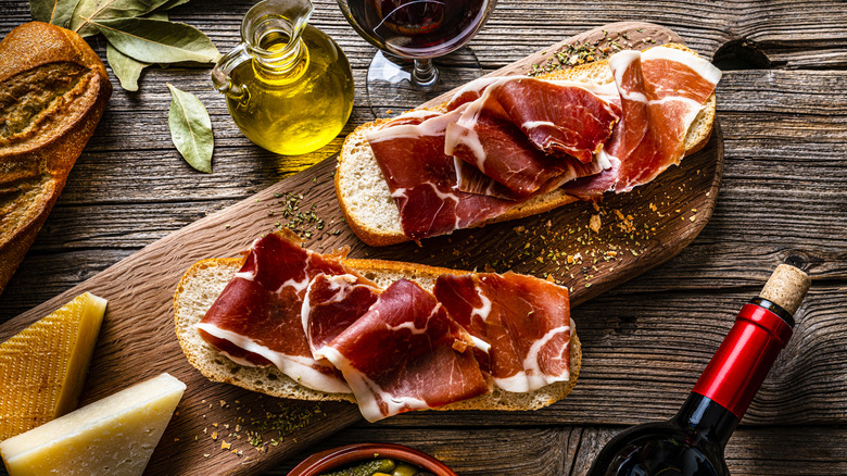A cutting board with sliced bread covered with jamón Iberíco, surrounded by Manchego cheese, olive oil, a bottle of wine, and a glass of wine.