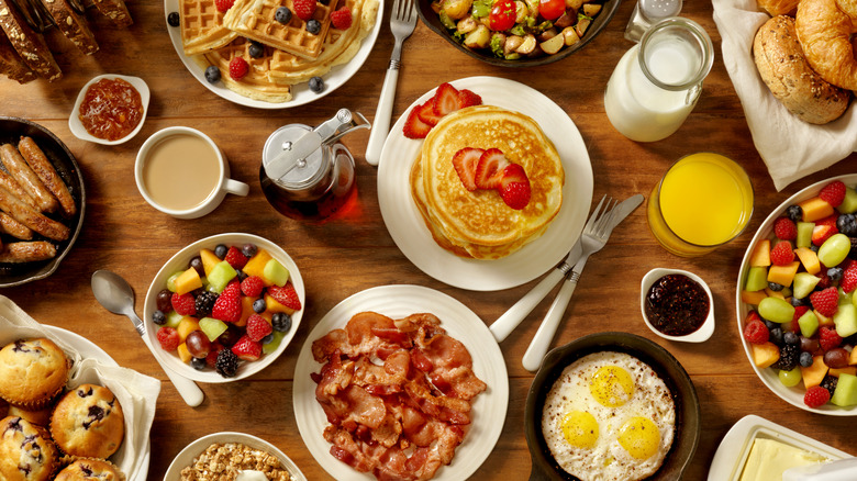 Top-down view of a wooden table with a variety of breakfast staples, including coffee, pancakes, fruit, bacon, eggs, waffles, and more