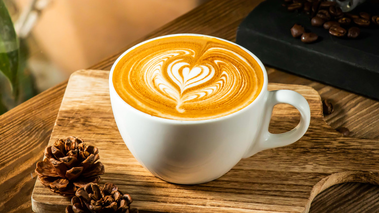 A latte in a white mug on a wooden board next to pine cones