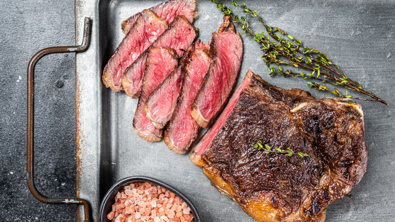 Cooked steak with several medium-rare slices cut on a metal tray with a spring of herbs and a small bowl of red, large-grained salt