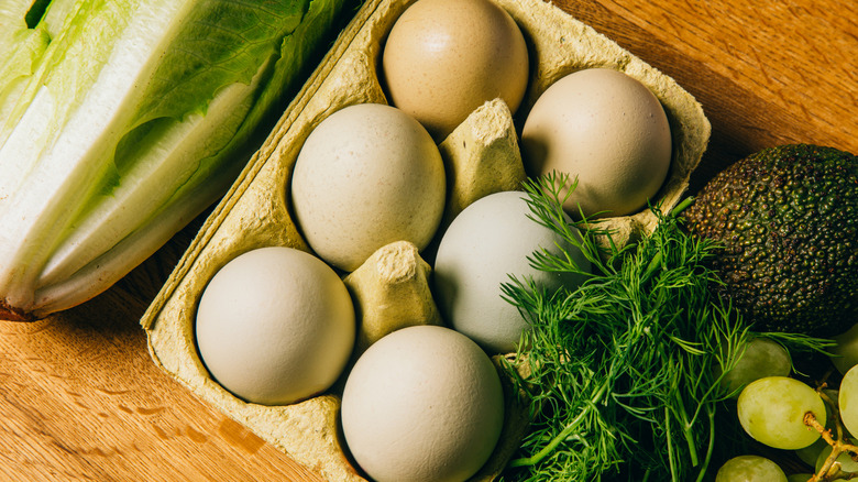 A half dozen of eggs sit in a carton next to green produce