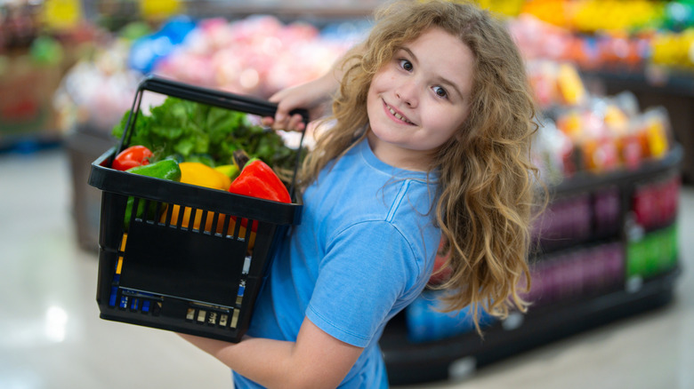 A smiling child holds a shopping basket full of vegetables in a supermarket produce section.