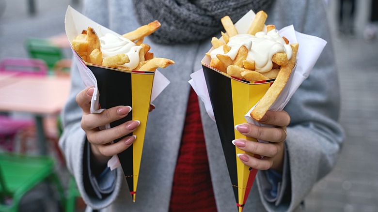 Person holding two paper cones filled with Belgian fries and topped with mayonnaise