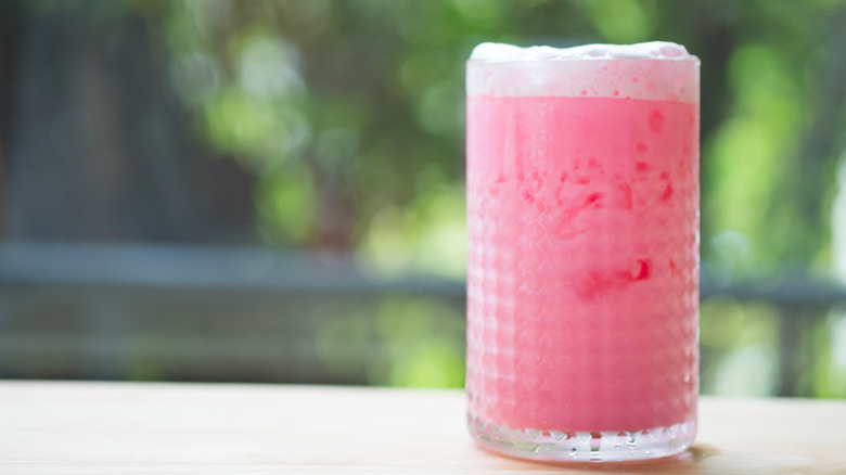 Layered pink drink in a clear glass with foam on top. On a white table, and blurred outdoorsy background.