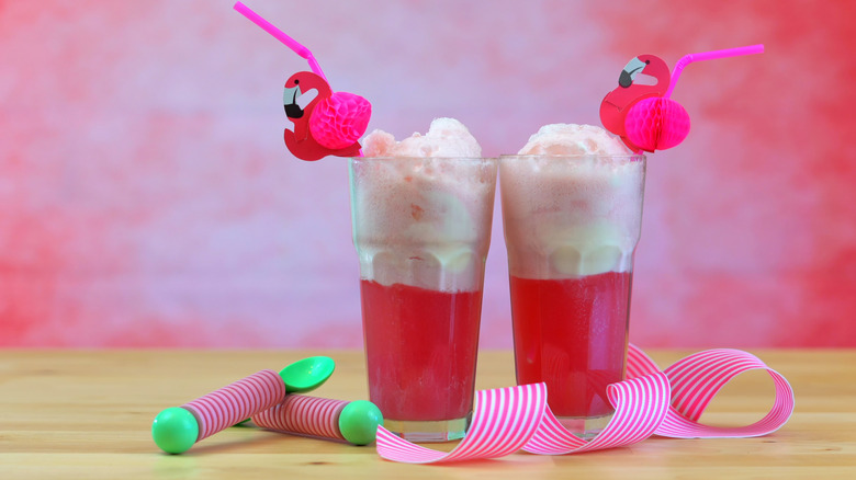 Two tall, clear glasses with pink cream soda. Each glass has a pink straw with flamingo decorations attached, and festive, striped spoons and paper around. All on a wooden table, with a texturized pink background.