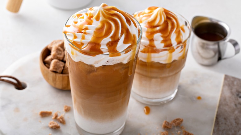 Two iced lattes topped with whipped cream and caramel sauce from an overhead view on a white marble table
