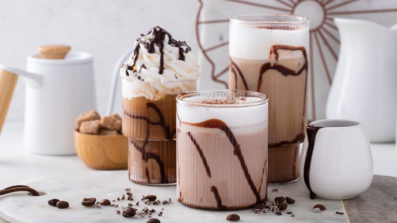 Chocolate frappes and coffees topped with either cold foam or whipped cream and chocolate syrup placed on a white marble table in different sized glasses