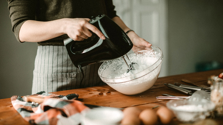 Woman whipping cream in a clear bowl with a hand mixer on a wooden table