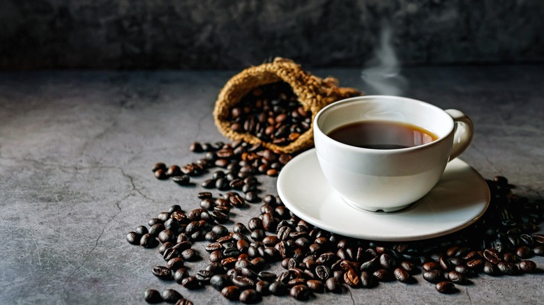 Cup of hot coffee in a white mug on a white saucer surrounded by espresso beans on a gray surface