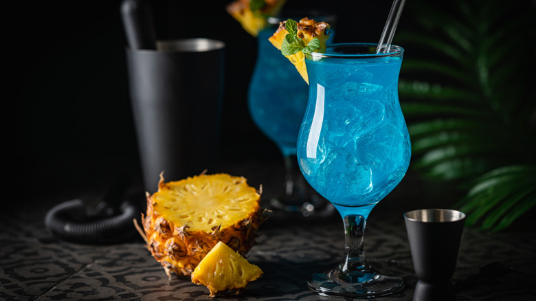 A blue cocktail in a hurricane glass garnished with a pineapple piece on a black patterned surface with a small cut bottom half of pineapple and a black jigger on either side and a black tall cup and another blue drink in the background