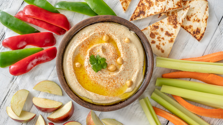 Hummus in a brown dish garnished with whole chickpeas and parsley, surrounding by veggie, apple, and pita slices