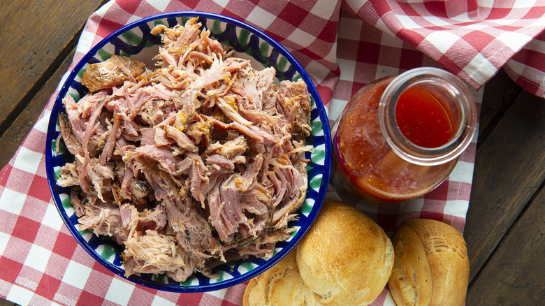 Shredded meat in a bowl alongside vinegar-based bbq sauce and rolls 