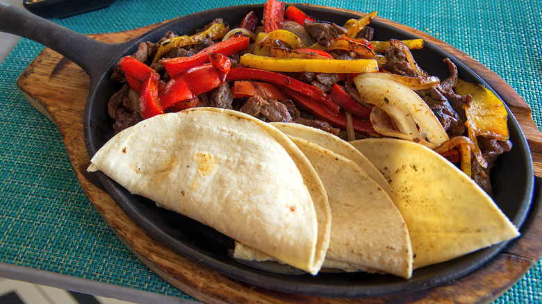 A hot fajita griddle with tortillas, meat, peppers, and onions on a wooden cutting board