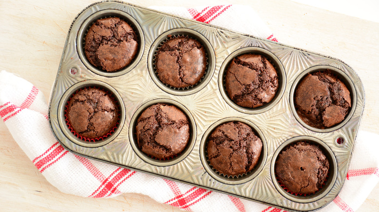 A batch of cooked brownies sit in a metal muffin tin against a red and white tea towel