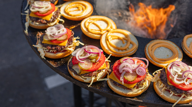 Open-faced burgers sitting around an open flame, piled high with toppings like tomatoes and onions