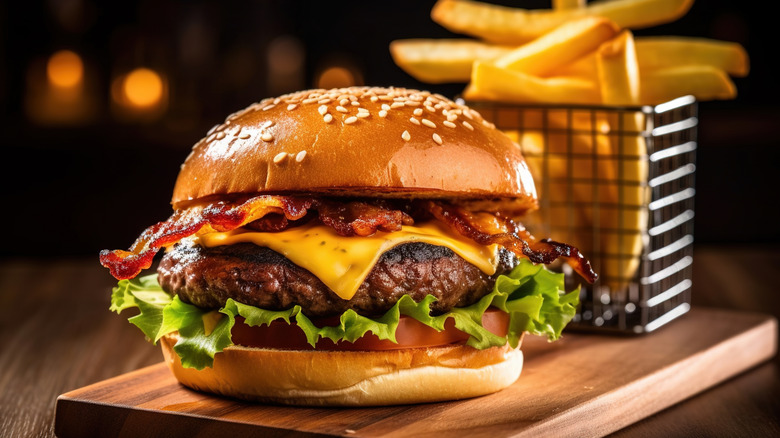 A burger with bacon, cheese, and lettuce with a sesame seed bun sits on a wooden cutting board