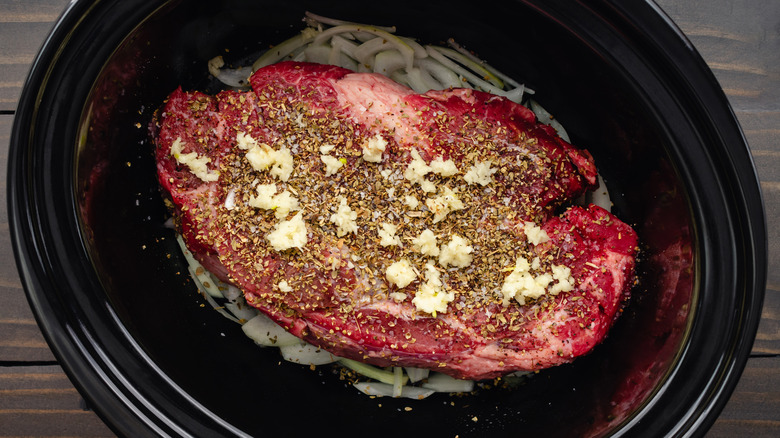 A beef chuck roast sits on a bed of aromatics in a classic crock pot.