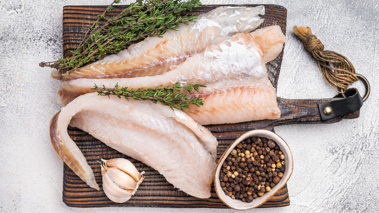 raw pollock on a wooden board surrounded by herbs and seasonings on a white background