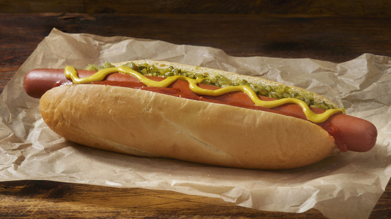 Extra-long hot dog in bun topped with relish, mustard, and ketchup on top of parchment paper