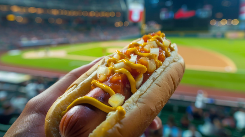 Hand holding a hot dog with baseball stadium in background