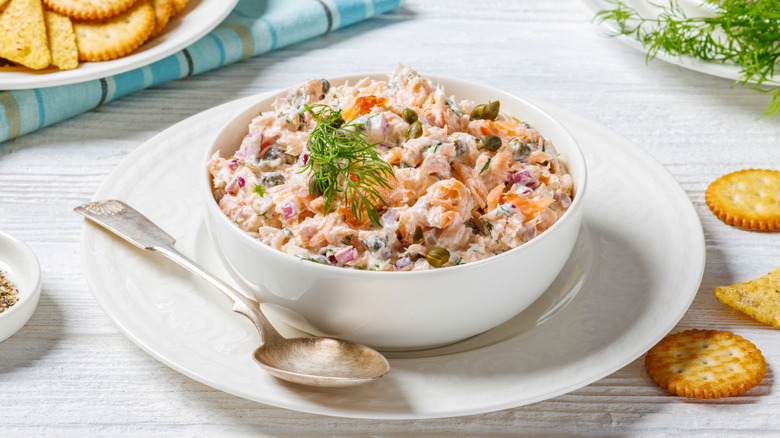 Salmon salad with fresh dill on top sits in a white bowl with crackers near it