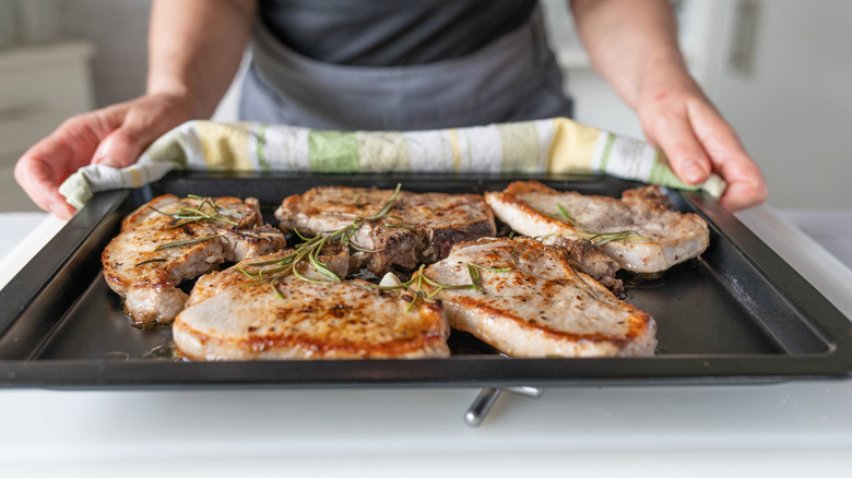 A sheet tray of pork chops topped with herbs held by a cook with a rag