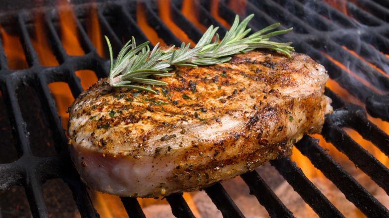 A grilled and heavily seasoned pork chop topped with a sprig of rosemary over the flames of a grill