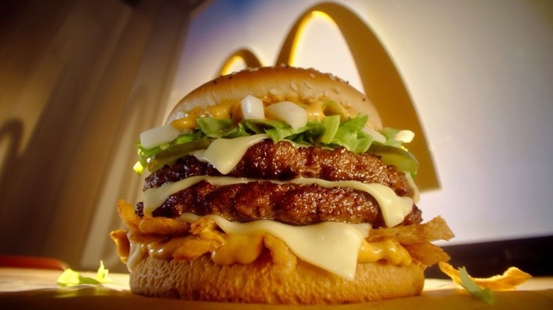The magnificent Big Arch sits on a table, dripping with sauce, cheese, lettuce, and crispy onions, in front of a window with a classic McDonald's M