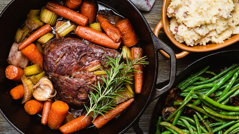 Pot roast displayed inside of a Dutch oven with roasted vegetables, fresh rosemary, and sides of mashed potatoes and green beans.