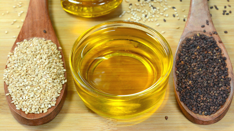 Two spoonfuls of seeds, one light-colored and one dark-colored, sit on wooden spoons next to a bowl of seed oil
