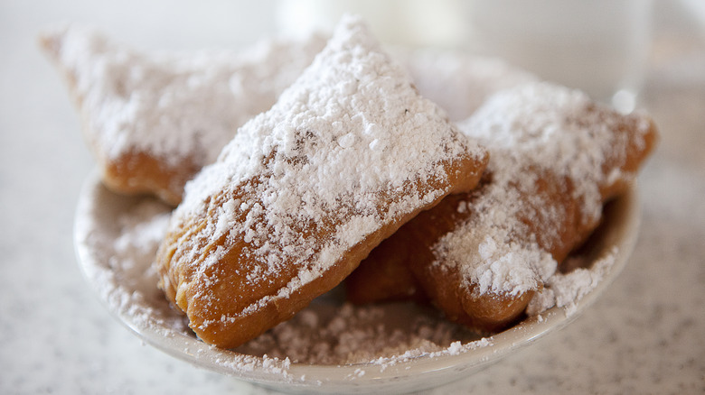 Close up of a pile of beignets covered in powdered sugar on a small plate. All on a white, speckled table.