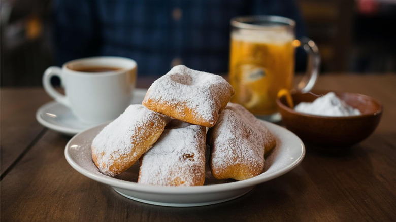 Pile of beignets on a small white plate, covered in powdered sugar on a wooden table. Two cups of coffee blurred in the background. One in a small white mug, the other in a tall glass mug, with a bowl of powdered sugar to the side.