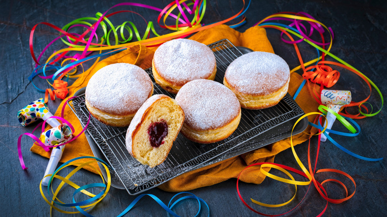 Tray of German doughnuts known as fasnachts with powdered sugar sprinkled on top, with on fasnacht cut open showing red jam filling. Party decorations are surrounding the tray on top of a yellow-orange napkin, on a gray table.