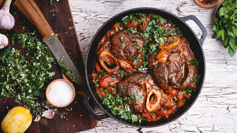 A cast iron pot of osso bucco next to a cutting board with garlic lemon herbs and a knife