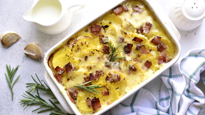 Potato casserole in a white dish surrounded by herbs, cloves of garlic, cream, and a salt shaker
