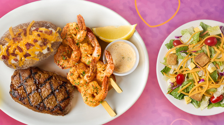 Plate with a small steak, skewered shrimp, and a loaded baked potato next to a salad.