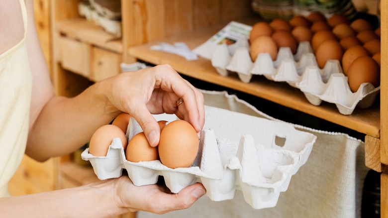 A woman grabs a brown egg from a cardboard carton, multiple dozens sit in cartons in the background