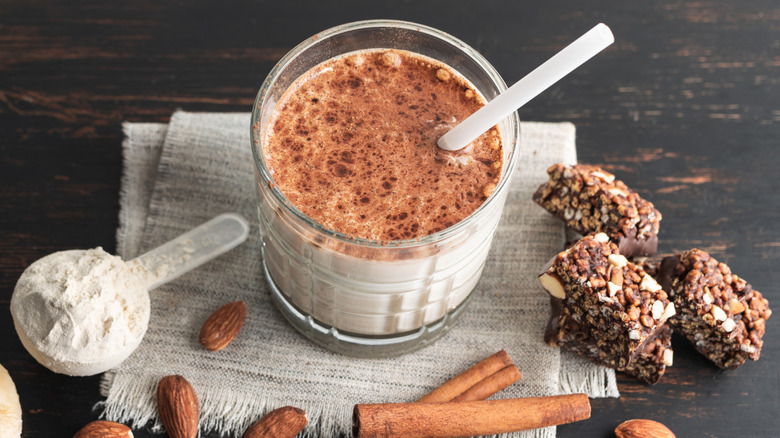 A cinnamon-topped protein shake with a straw, granola, nuts, and a scoop of protein