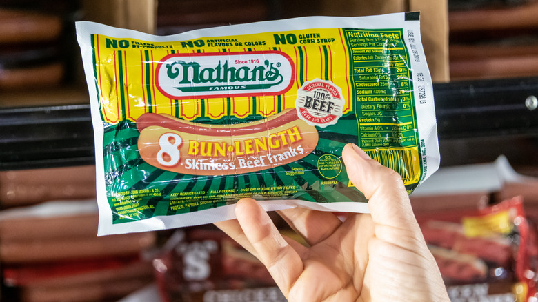 Shoppers hand holding a package of Nathan's brand bun length skinless beef franks in a supermarket aisle