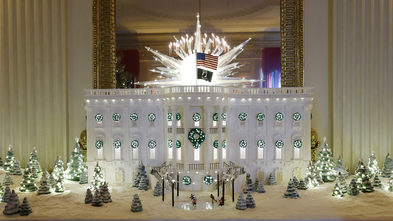 gingerbread remake of the White House with two flags at the top and several decorative wreaths and snow-covered evergreen trees on a snowy lawn